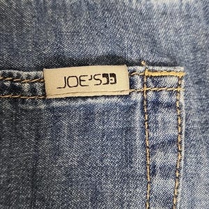 May include: Blue denim fabric featuring a rectangular beige fabric tag with the text JOE'S printed in black. The denim has visible gold-toned stitching along the seams and pocket edge, showcasing the texture of the woven cotton material.
