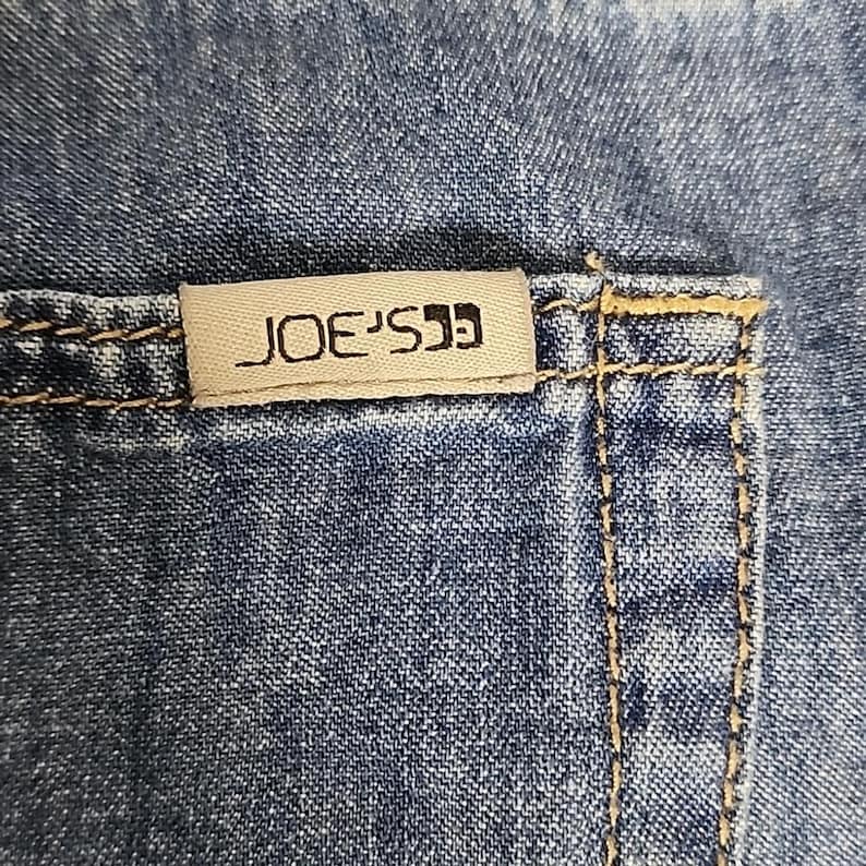 May include: Blue denim fabric featuring a rectangular beige fabric tag with the text JOE'S printed in black. The denim has visible gold-toned stitching along the seams and pocket edge, showcasing the texture of the woven cotton material.