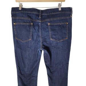 May include: Dark wash denim jeans with two back pockets and a button closure. The jeans are hanging on a wooden hanger against a white background. The jeans have a classic five-pocket design.