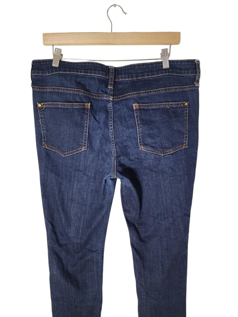 May include: Dark wash denim jeans with two back pockets and a button closure. The jeans are hanging on a wooden hanger against a white background. The jeans have a classic five-pocket design.