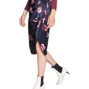 May include: A navy blue midi skirt with a floral pattern in shades of pink and red. The skirt has a wrap design with a ruffled detail. The model is wearing white ankle boots with a small heel.