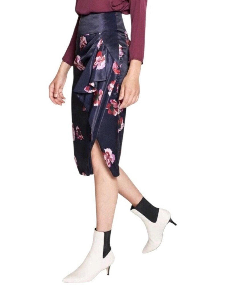 May include: A navy blue midi skirt with a floral pattern in shades of pink and red. The skirt has a wrap design with a ruffled detail. The model is wearing white ankle boots with a small heel.