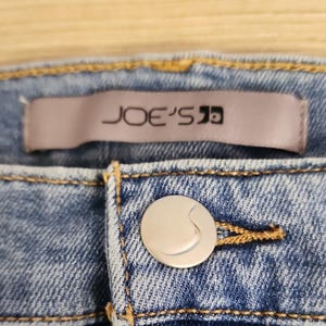 May include: Close-up of light blue denim jeans with a silver button and the brand name "JOE'S" on a label. The jeans have yellow stitching and a button closure.