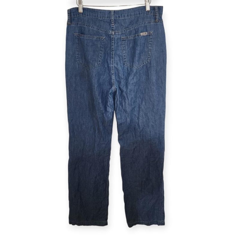 May include: A pair of medium-wash blue denim jeans featuring a high-waisted design, a button and zip fly closure, two back patch pockets, and a small rectangular metal brand tag attached to the upper right back pocket area.