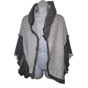 Op de afbeelding: Een grijze en donkergrijze poncho-stijl trui met capuchon. Het kledingstuk met open voorkant heeft een gefranjerde rand en donkergrijze mouwen. De trui wordt op een mannequin getoond.