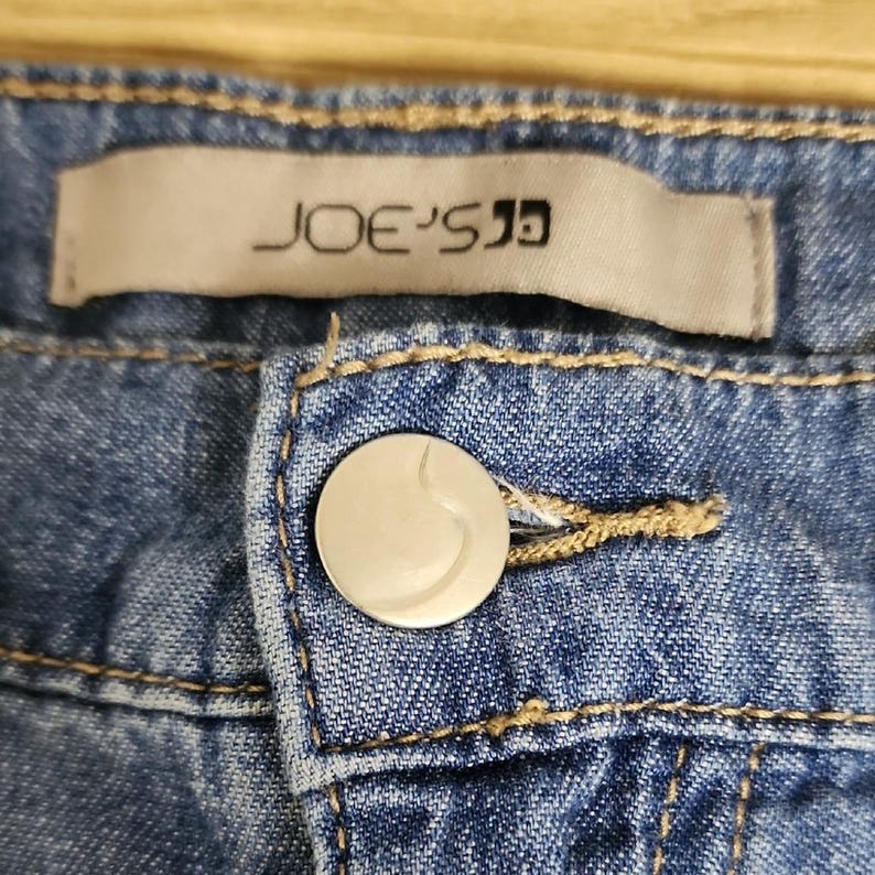 May include: Blue denim jeans featuring a silver-toned metal button closure and a rectangular fabric label stitched above the fly. The label displays the text JOE'S in black lettering. The denim fabric shows visible stitching in a tan thread colour.