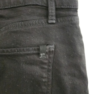 May include: Close-up of black denim jeans. The image shows the back pocket, belt loop, and stitching details. A small black label is attached to the back pocket.