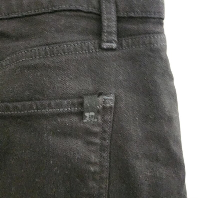 May include: Close-up of black denim jeans. The image shows the back pocket, belt loop, and stitching details. A small black label is attached to the back pocket.