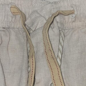 May include: Close-up of the waistband of a pair of white linen pants with a beige drawstring. The drawstring has red stitching.