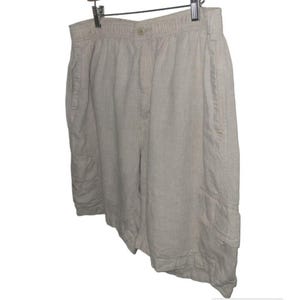 May include: Beige linen shorts with an elastic waistband and button closure. The shorts have side pockets and a cargo pocket on the left leg. The shorts are hanging on a metal hanger.