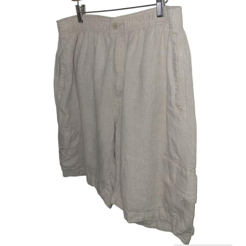 May include: Beige linen shorts with an elastic waistband and button closure. The shorts have side pockets and a cargo pocket on the left leg. The shorts are hanging on a metal hanger.