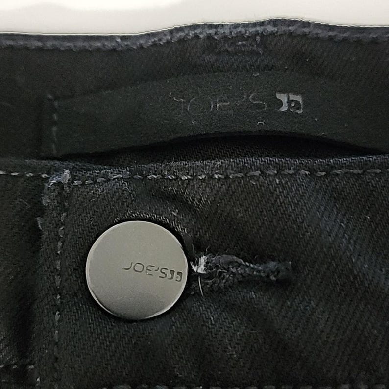 May include: Close-up of black denim fabric with a silver button. The button is embossed with the text "JOE'S" and the brand logo. The stitching and fabric texture are visible.