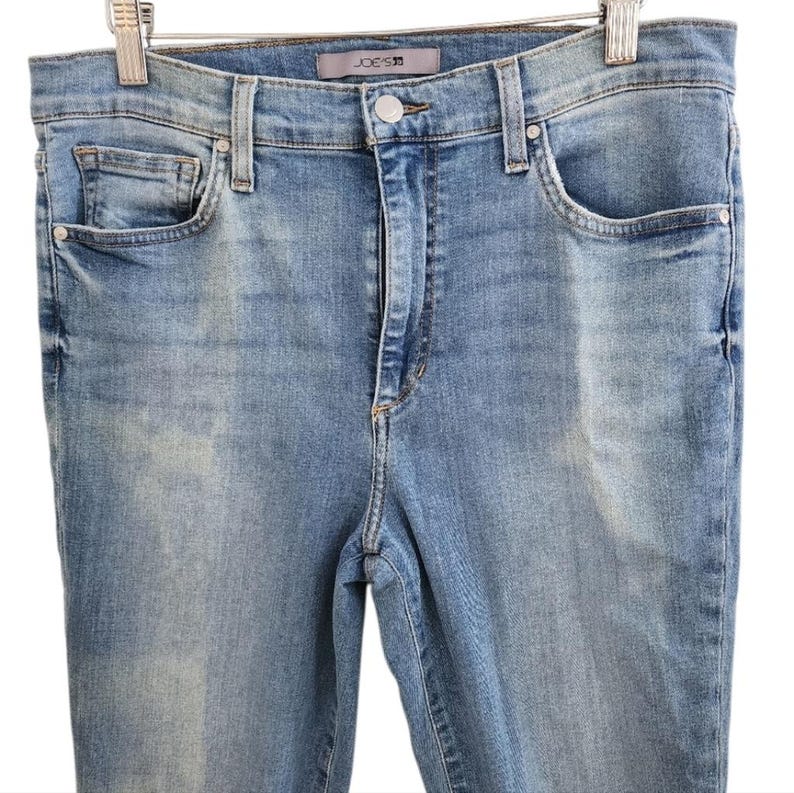 May include: Light blue denim jeans with a classic five-pocket design. The jeans feature a button and zipper closure, belt loops, and the brand name "JOE'S" on the waistband. The jeans are hanging on a metal hanger.