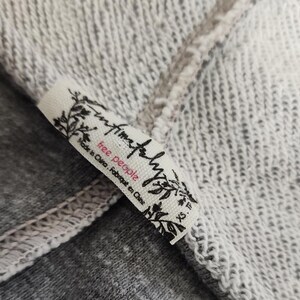 Op de afbeelding: Close-up van een wit stoffen label met de woorden "free people" in roze en zwarte letters. Het label is bevestigd aan een grijs en wit gem&ecirc;leerde stof. Het label leest ook "Made in China. Fabriqu&eacute; en Chine" en "XS. TP."