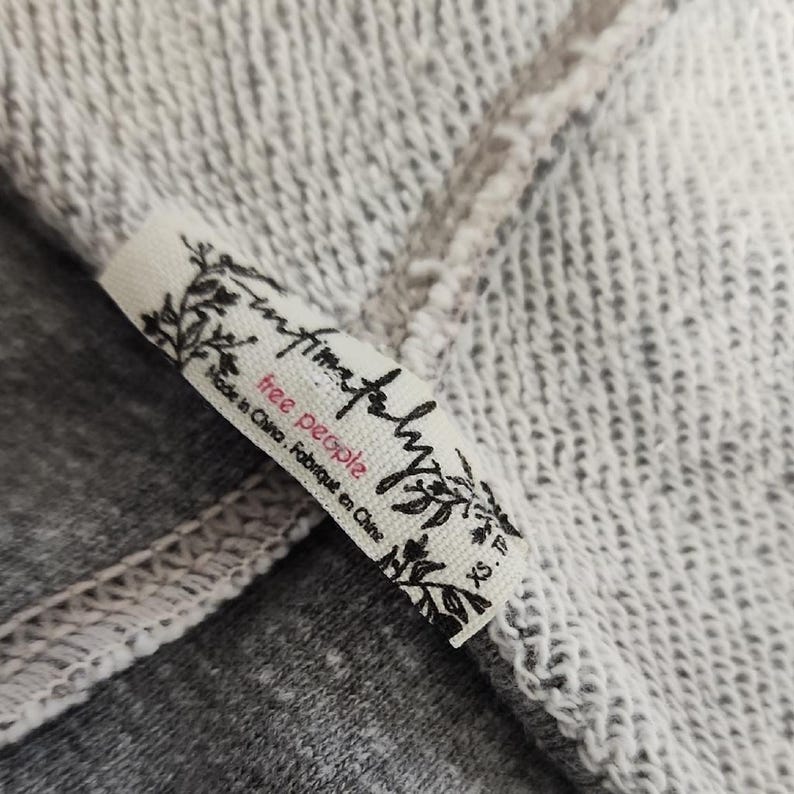Op de afbeelding: Close-up van een wit stoffen label met de woorden "free people" in roze en zwarte letters. Het label is bevestigd aan een grijs en wit gem&ecirc;leerde stof. Het label leest ook "Made in China. Fabriqu&eacute; en Chine" en "XS. TP."