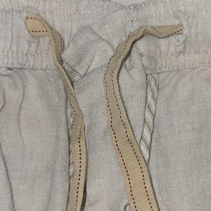 May include: Close-up of light gray linen shorts with a beige drawstring. The drawstring has a red stitched pattern. The shorts have a textured appearance and a relaxed fit, suggesting comfort and casual wear.