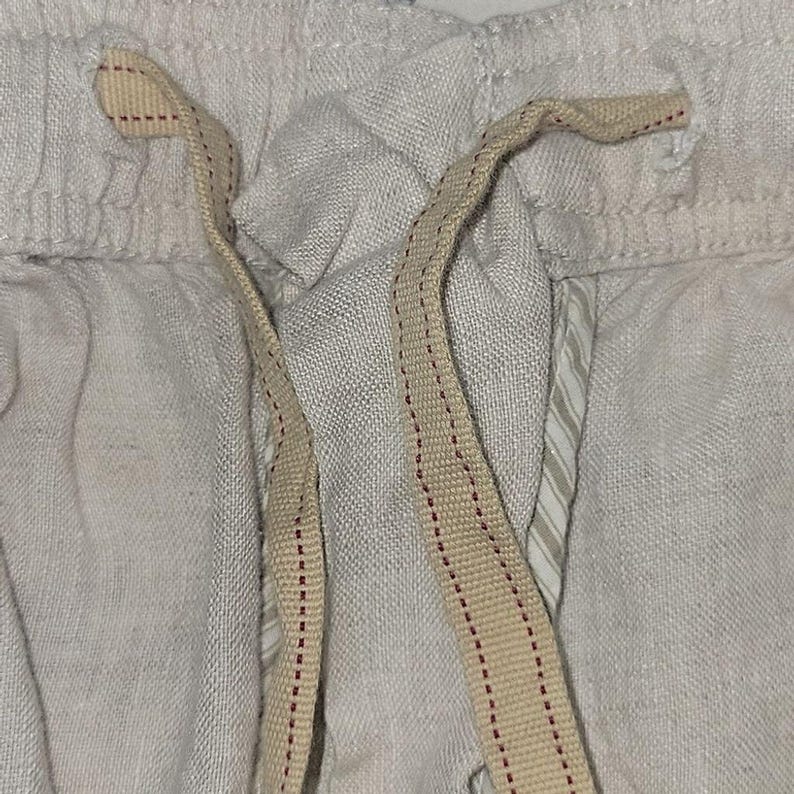 May include: Close-up of light gray linen shorts with a beige drawstring. The drawstring has a red stitched pattern. The shorts have a textured appearance and a relaxed fit, suggesting comfort and casual wear.