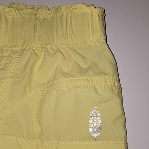 May include: Close-up of yellow shorts with an elasticated waistband. The shorts feature a silver logo on the lower left. The fabric has visible stitching and a slightly wrinkled texture.