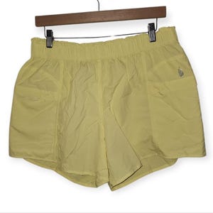 May include: Pale yellow athletic shorts with an elasticated waistband and two side pockets. The shorts are hanging on a wooden hanger. A small logo is visible on the right side. The shorts are made of a lightweight, slightly shiny material.