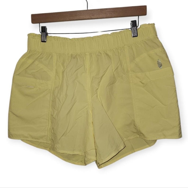 May include: Pale yellow athletic shorts with an elasticated waistband and two side pockets. The shorts are hanging on a wooden hanger. A small logo is visible on the right side. The shorts are made of a lightweight, slightly shiny material.