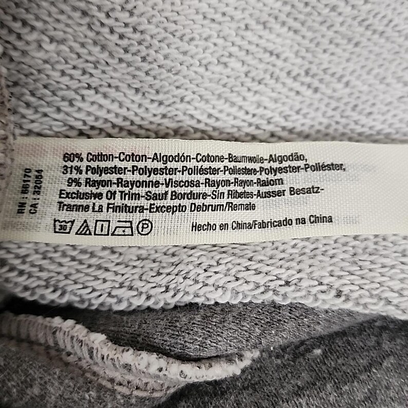 Op de afbeelding: Een close-up van een kledinglabel met tekst over de samenstelling van de stof: 60% katoen, 31% polyester en 9% rayon. Het label bevat ook wasvoorschriften en de tekst "Hecho en China/Fabricado na China."