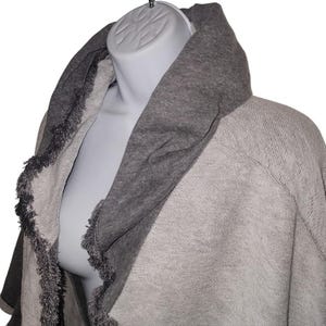 Op de afbeelding: Een grijze sjaal-stijl trui met een brede, donkergrijze kraag. De randen van de trui zijn afgewerkt met een franje detail. De trui wordt getoond op een mannequin.