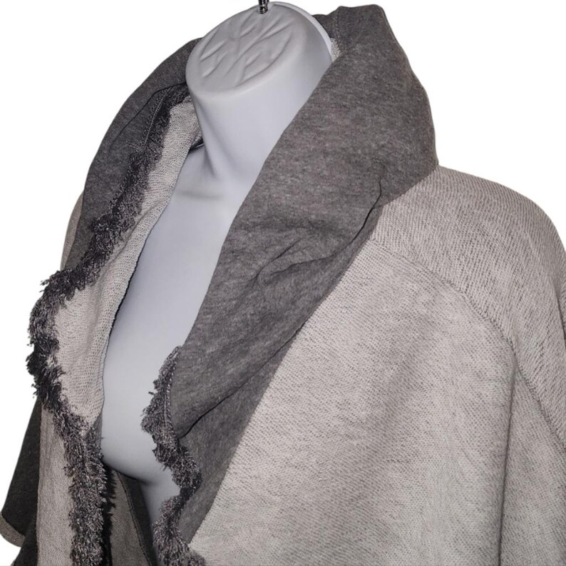 Op de afbeelding: Een grijze sjaal-stijl trui met een brede, donkergrijze kraag. De randen van de trui zijn afgewerkt met een franje detail. De trui wordt getoond op een mannequin.