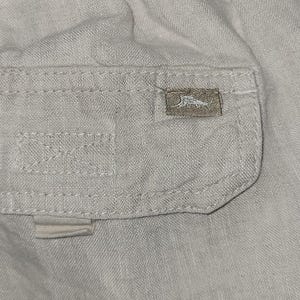 May include: A close-up of a light beige cargo pocket with a small brown patch that has a bird logo on it.