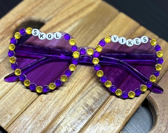 May include: Purple heart-shaped sunglasses with yellow rhinestones around the frame. The sunglasses have the words "SKOL" and "VIKES" spelled out in white letters on the frames.
