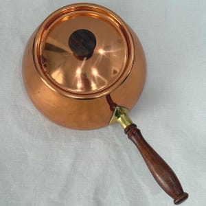 May include: A round copper pot with a polished finish, featuring a matching lid with a dark brown wooden knob. The pot includes a long, slender handle made of dark brown wood, attached to the body with a bright brass-colored metal fitting.