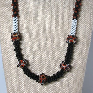 May include: A beaded necklace featuring a long strand of small matte black beads interspersed with segments of white beads and five larger, round, patterned focal beads in shades of orange, black, and white with a distinct eye-like design.