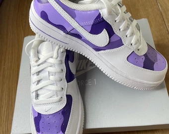 May include: A pair of white Nike Air Force 1 sneakers with a purple and light purple camouflage pattern. The shoes have a white swoosh logo and the text "Nike" and "Force 1" on the side.