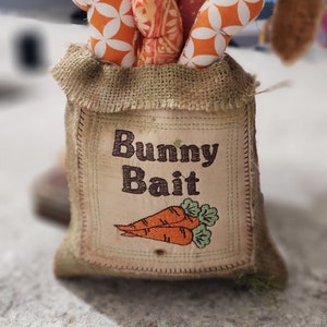 Könnte beinhalten: Ein Jutesack mit der Aufschrift "Bunny Bait", die darauf gestickt ist. Der Sack ist mit orangefarbenen und weißen Stoffkarotten gefüllt.
