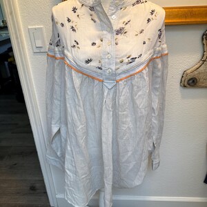 May include: A white and blue striped long-sleeved shirt with a floral print on the top portion. The shirt has a button-up front and a mandarin collar.