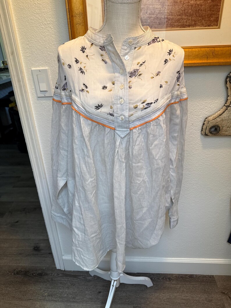 May include: A white and blue striped long-sleeved shirt with a floral print on the top portion. The shirt has a button-up front and a mandarin collar.