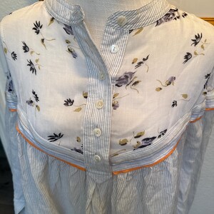 May include: A white button-down shirt with a floral print on the top half and thin blue stripes on the bottom half. The shirt has a mandarin collar and a single button closure.