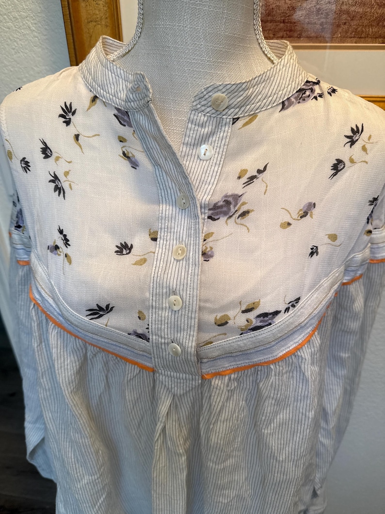 May include: A white button-down shirt with a floral print on the top half and thin blue stripes on the bottom half. The shirt has a mandarin collar and a single button closure.