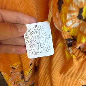 May include: A close-up of a white tag with the text "Free People" written in black cursive script. The tag is attached to a bright orange garment with a floral pattern.