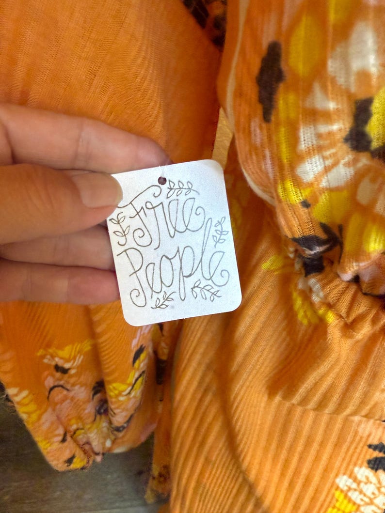 May include: A close-up of a white tag with the text "Free People" written in black cursive script. The tag is attached to a bright orange garment with a floral pattern.