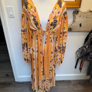May include: A long, orange and white striped maxi dress with a floral print. The dress has a deep V-neckline and long sleeves. The dress is cinched at the waist with a tie.