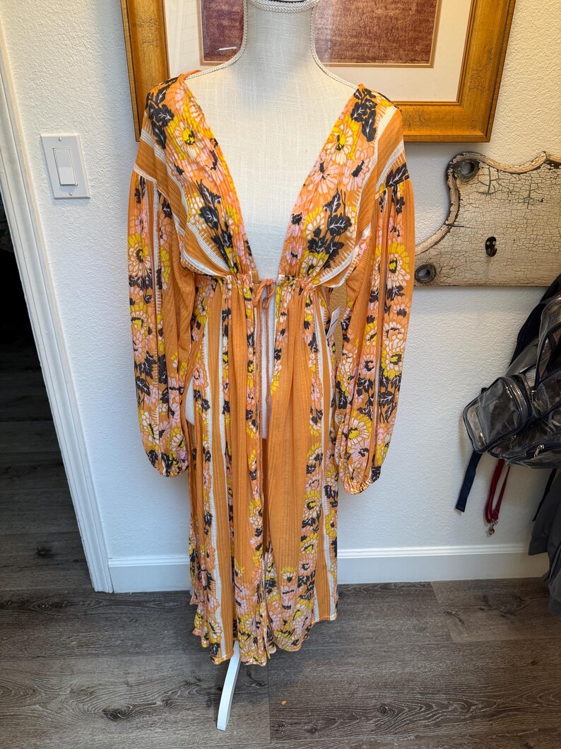 May include: A long, orange and white striped maxi dress with a floral print. The dress has a deep V-neckline and long sleeves. The dress is cinched at the waist with a tie.