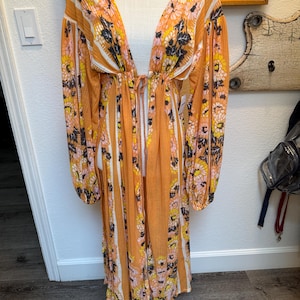 May include: A long, orange and white striped maxi dress with a floral print. The dress has a deep V-neckline and long sleeves. The dress is cinched at the waist with a tie.