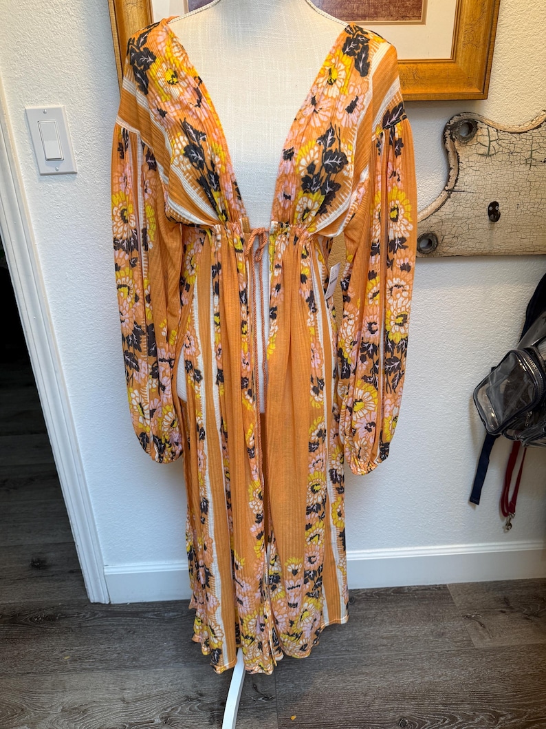 May include: A long, orange and white striped maxi dress with a floral print. The dress has a deep V-neckline and long sleeves. The dress is cinched at the waist with a tie.