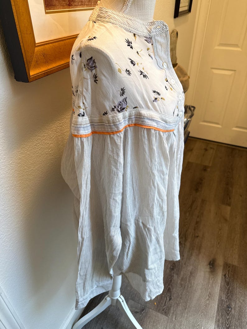 May include: A white and blue striped long-sleeved shirt with a floral print on the top half. The shirt has a button-up collar and a button closure.