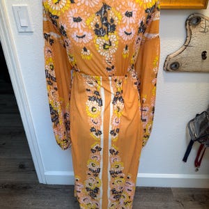 May include: A long, orange maxi dress with a floral print. The dress has a V-neckline, long sleeves, and a fitted waist. The floral print features pink, yellow, and black flowers on a light orange background.