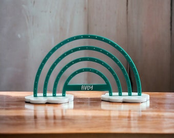 May include: A teal rainbow-shaped earring holder with the name "AVEY" printed on the front. The holder has four arched tiers with small holes for earrings, and sits on two white cloud-shaped bases. The background is a light brown wooden surface.