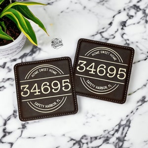 May include: Two brown leather coasters with stitched edges. Each coaster features a gold circle with the text "HOME SWEET HOME" and the number "34695" in gold. The text "SAFETY HARBOR, FL" is also in gold below the number.