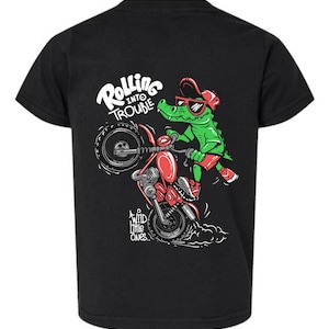 May include: A black short-sleeve t-shirt featuring a graphic of a green alligator wearing red sunglasses and a red cap, riding a red motorcycle on its back wheel. The text reads "ROLLING INTO TROUBLE" and "WILD LITTLE ONES" in white lettering.