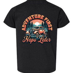 May include: A black short-sleeve t-shirt featuring a graphic print of a vintage orange car parked in front of a forest and sunset. The text ADVENTURE FIRST is arched above the car, and the words Naps Later are printed in script below the car.