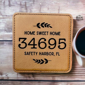 May include: A brown leather coaster with the text "Home Sweet Home", the house number "34695", and "Safety Harbor, FL" in black lettering. The coaster has a decorative leaf design at the top and bottom.